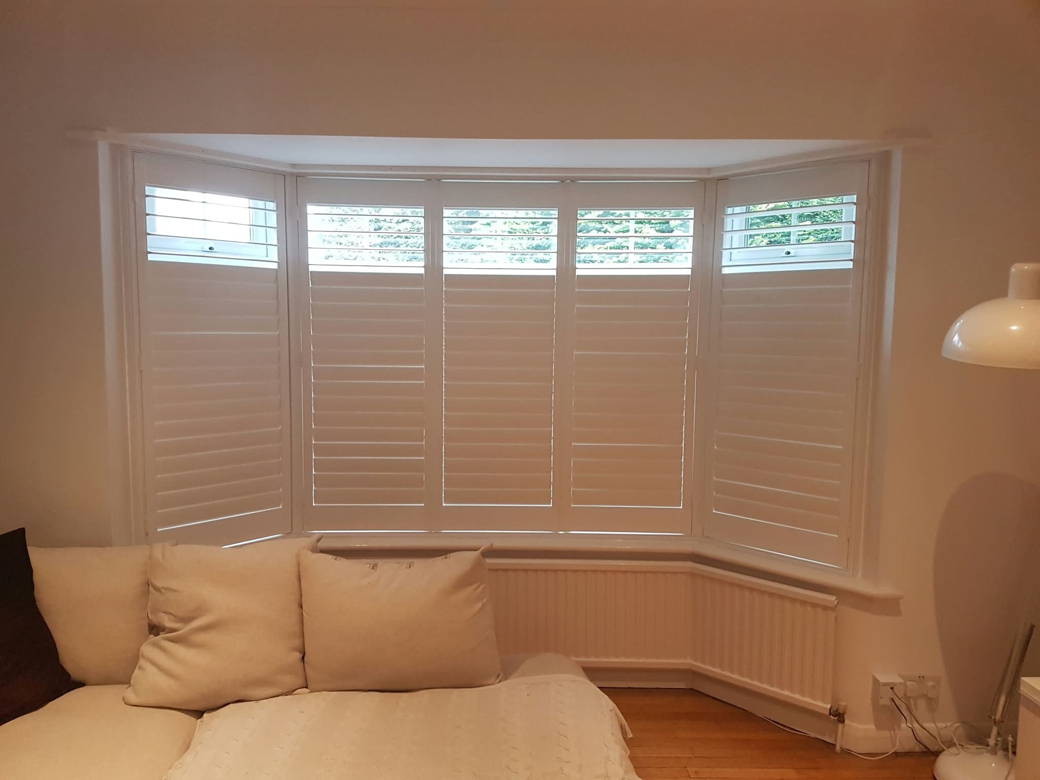 Full-height white shutters on a bay window living room — customer installation by Sustainable Shutters