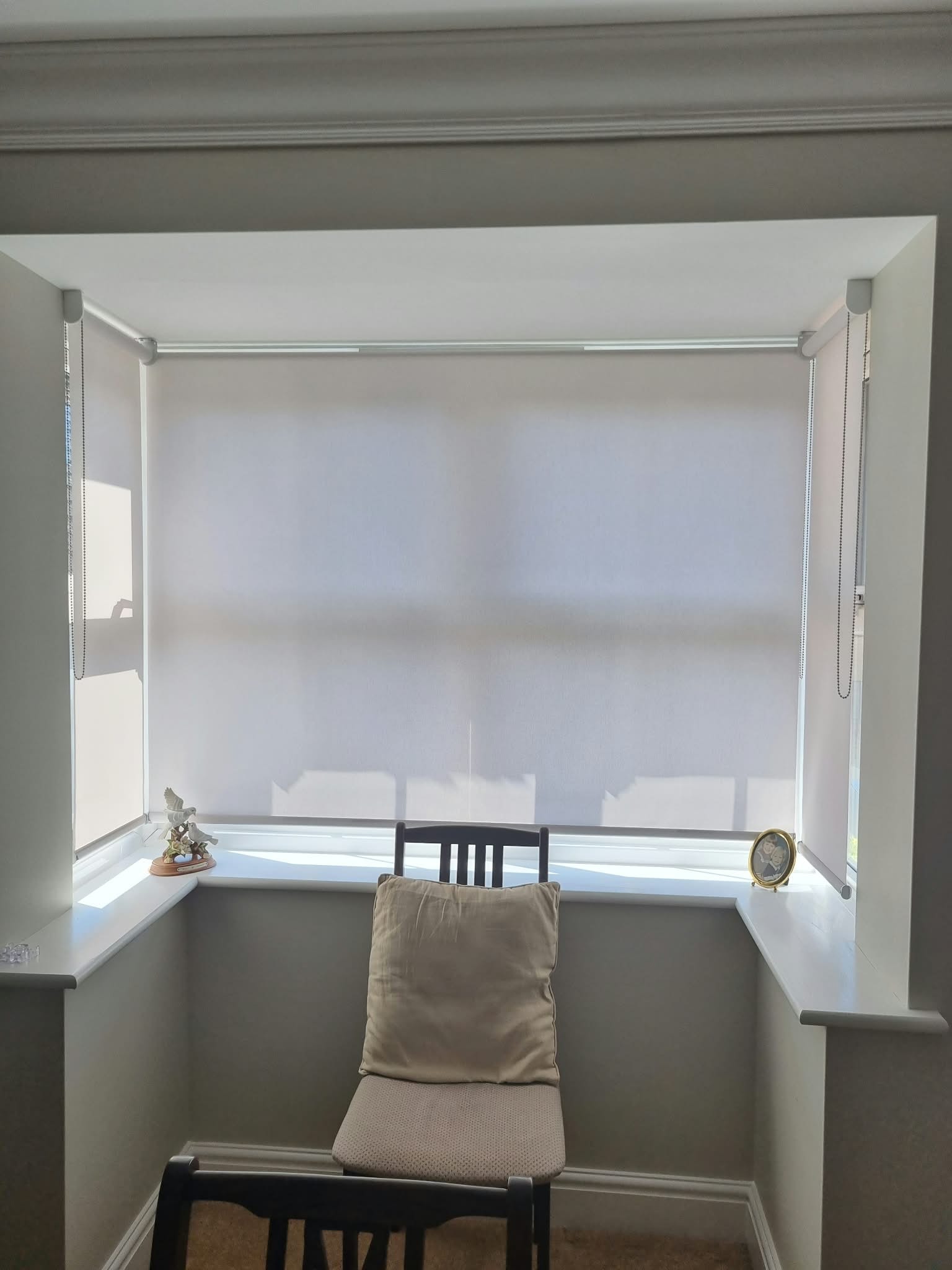 White roller blinds on a bay window with chair — Sustainable Shutters & Blinds
